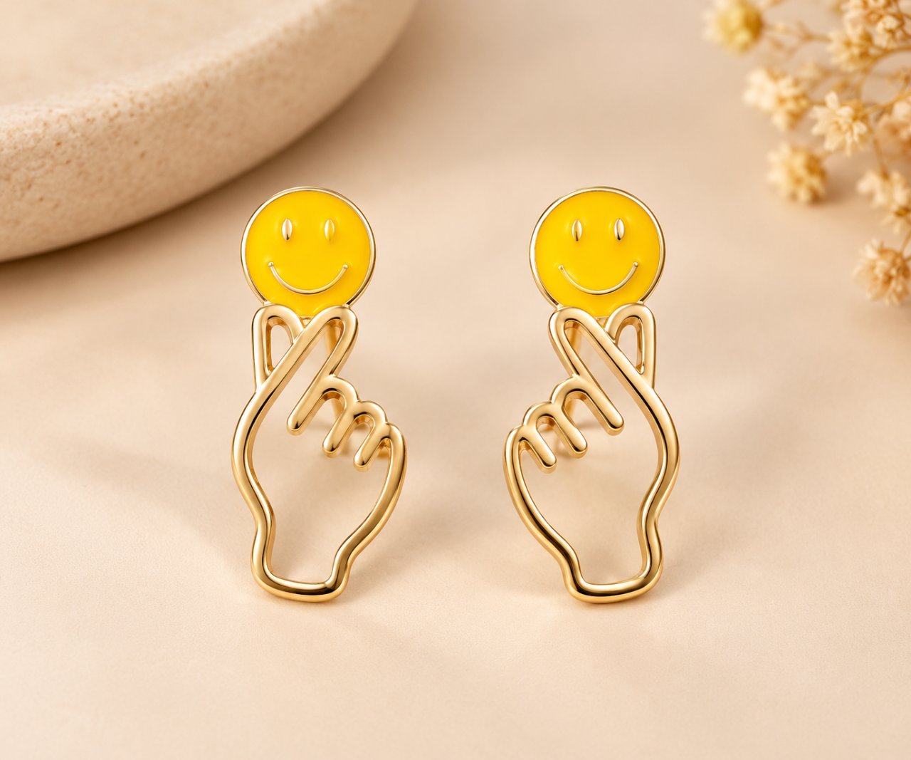Par de brincos em tom dourado sobre fundo bege, com carinhas amarelas sorridentes e mãos vazadas fazendo corações com os dedos.